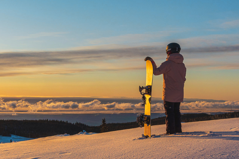 Personne en tenue de ski regarde un coucher de soleil avec un snowboard.