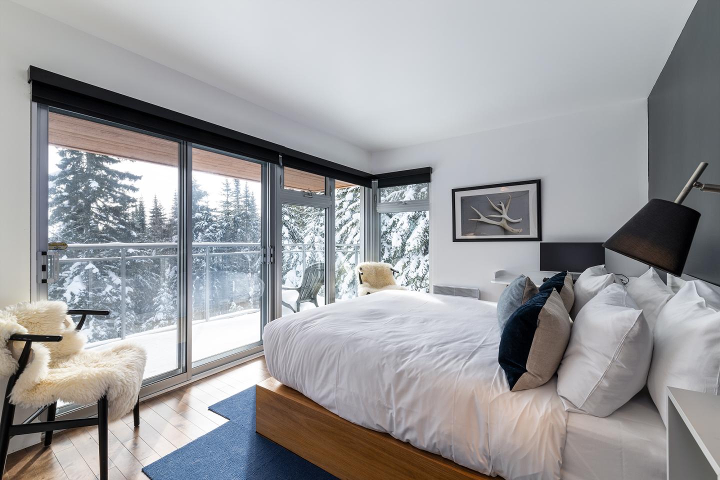 Chambre lumineuse avec lit blanc, fenêtres sur paysage enneigé.