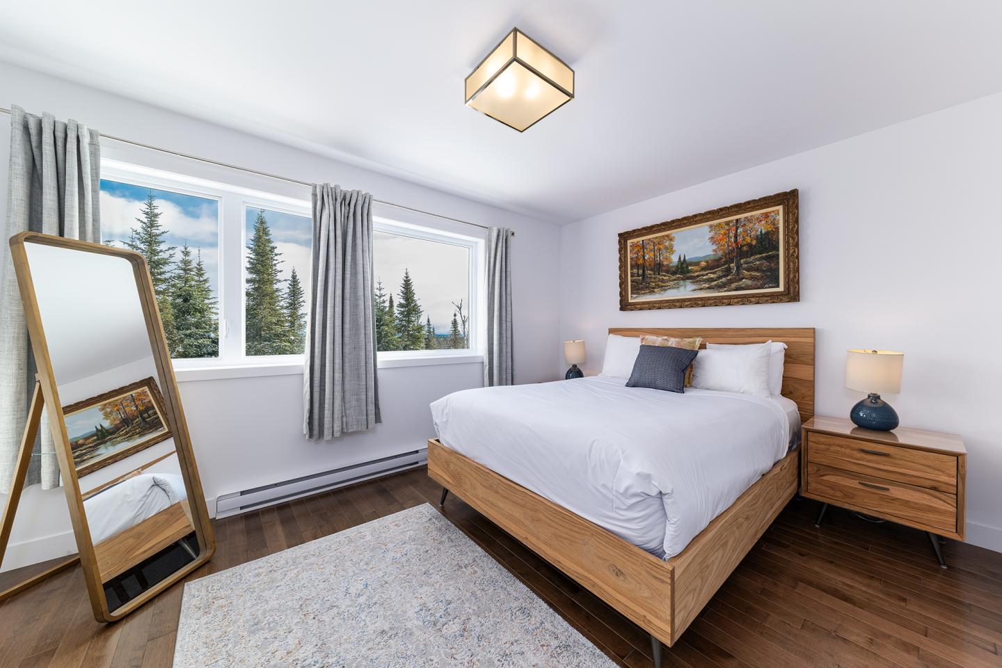 Chambre lumineuse avec lit double, miroir et tableau. Fenêtre avec vue sur les arbres.