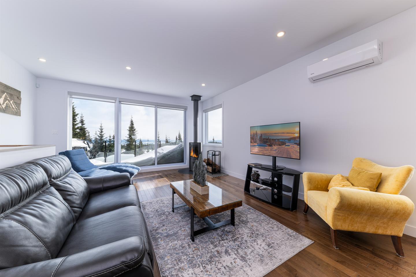 Salon moderne avec canapé noir, fauteuil jaune, table basse et vue sur la neige par la fenêtre.