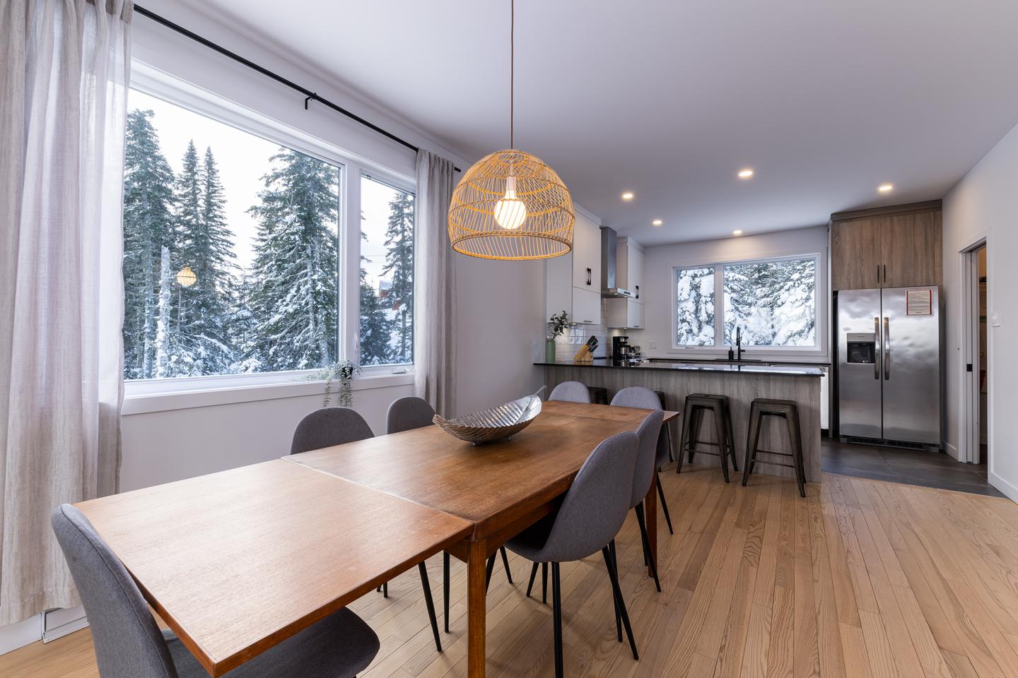 Salle à manger moderne avec vue sur la neige, table en bois, chaises grises, cuisine ouverte.