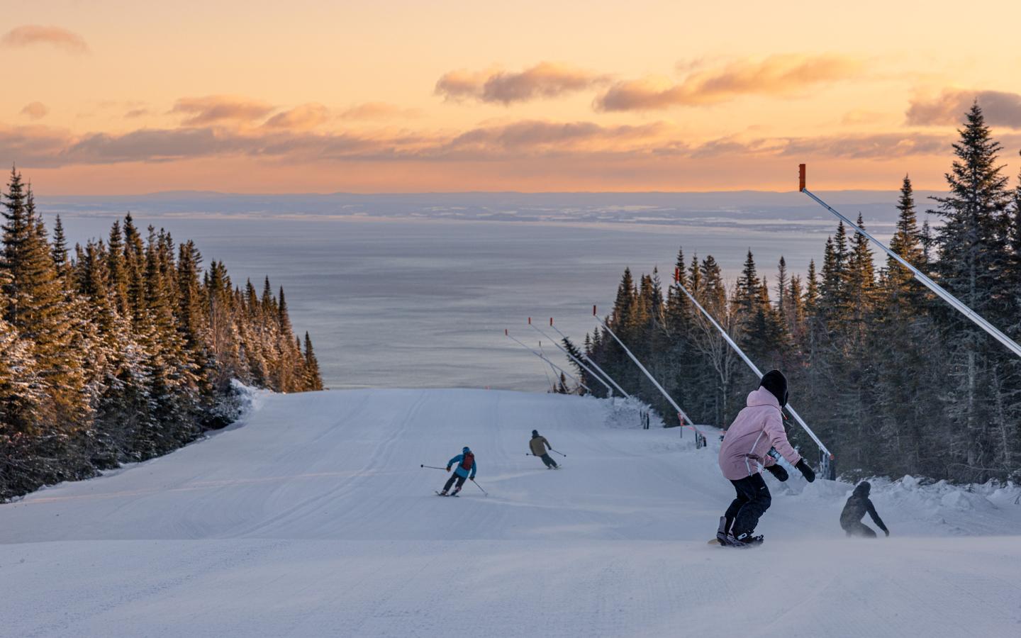 Skieurs sur une piste enneigée au coucher du soleil, entourés de sapins.