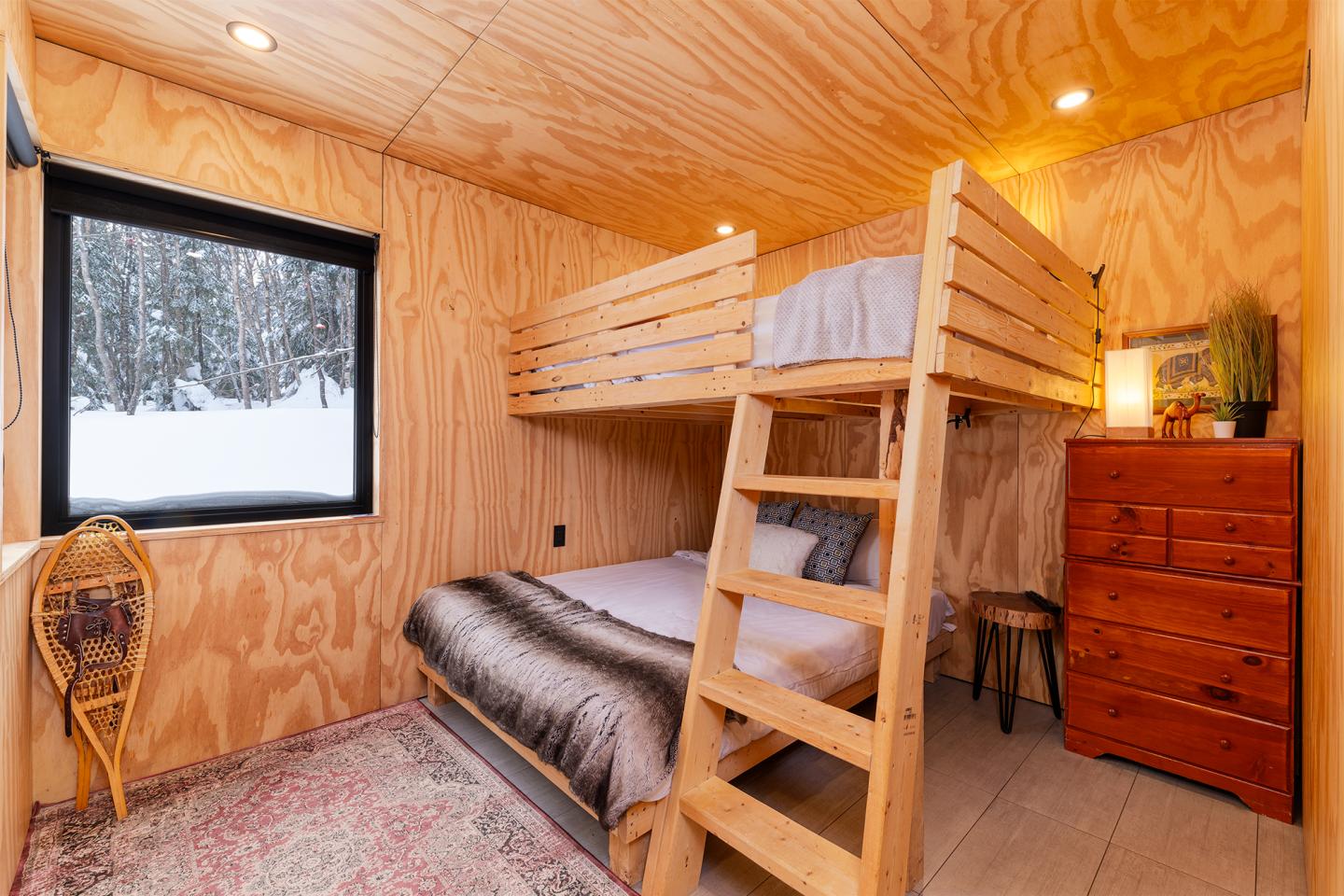 Chambre en bois avec lits superposés, fenêtre sur la neige, commode et lampe.