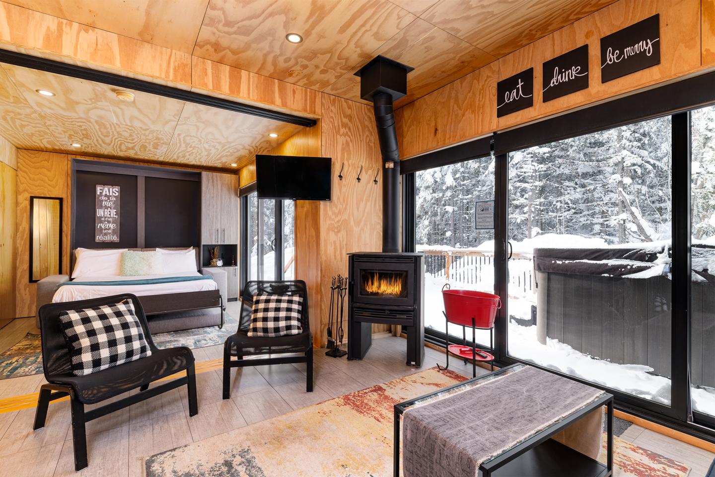 Intérieur d'une cabine moderne en bois avec poêle, fauteuils, et vue sur la neige.