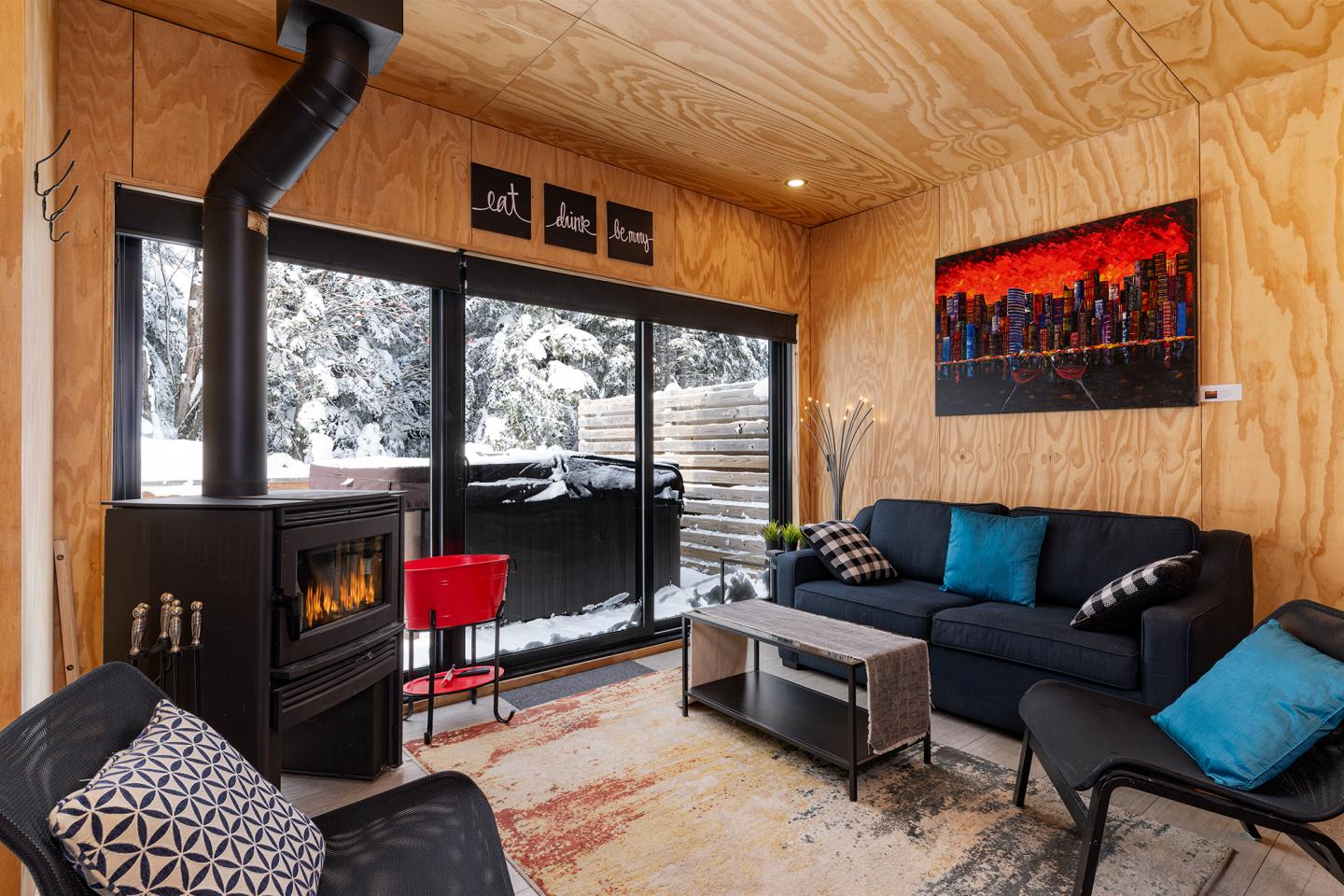 Salon chaleureux avec canapé bleu, poêle à bois et vues sur la neige à travers de grandes fenêtres.