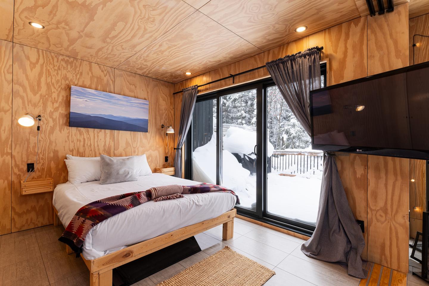 Chambre en bois avec lit, grandes fenêtres donnant sur la neige.