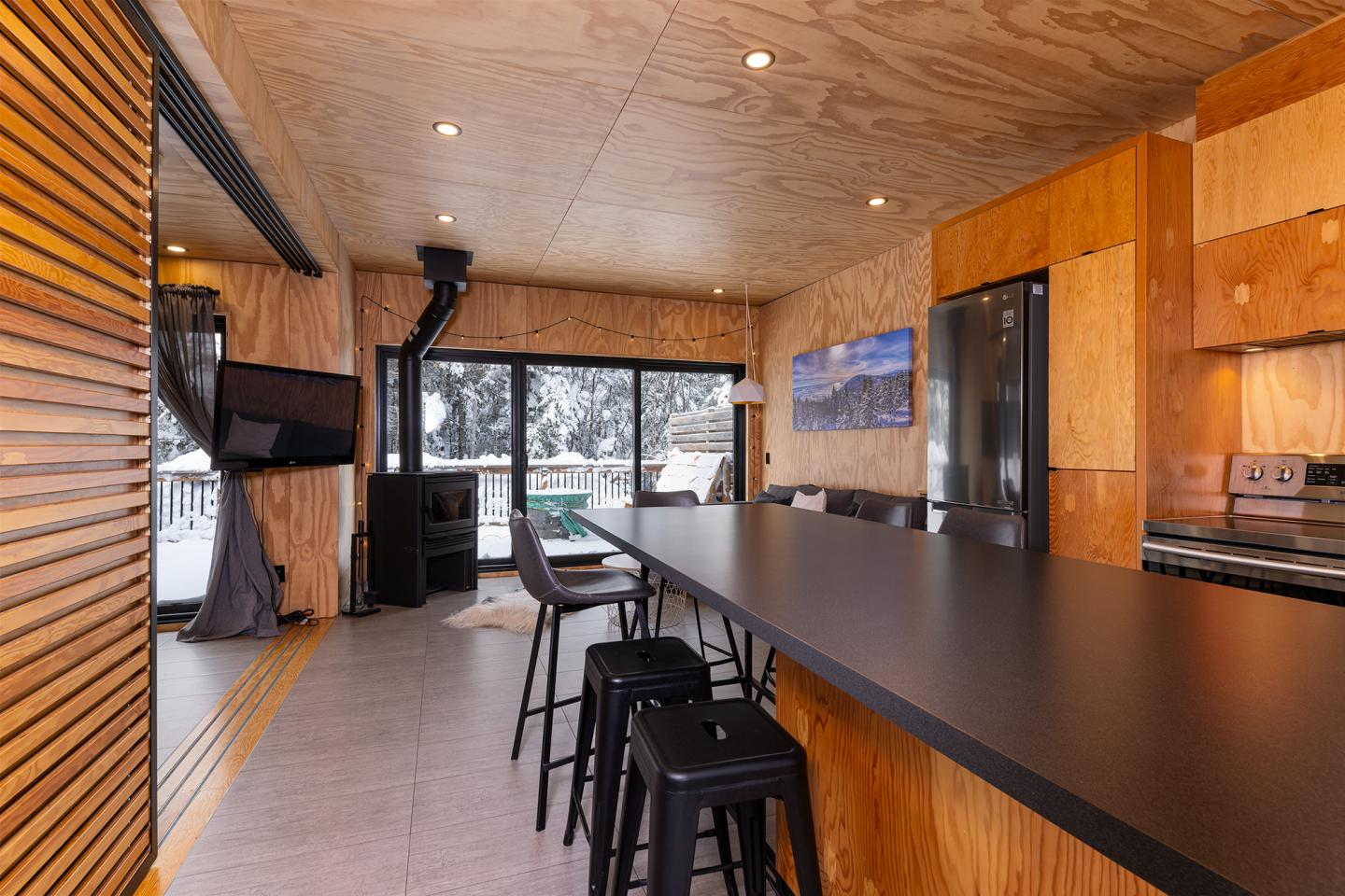Cuisine moderne en bois avec comptoir noir, chaises hautes et vue sur l'extérieur enneigé.