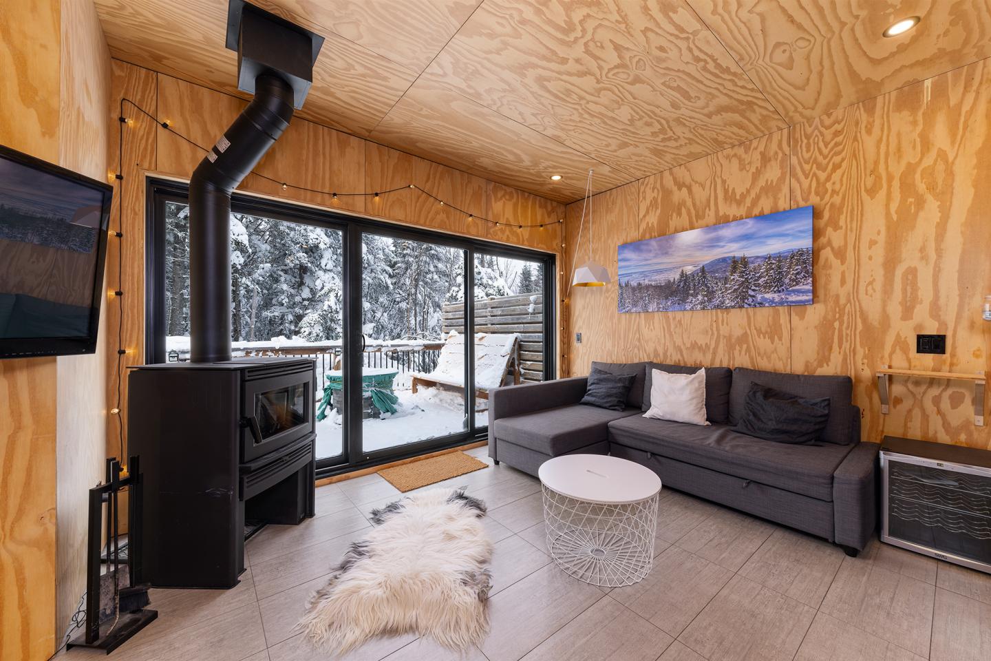 Salon cosy en bois, cheminée noire, canapé gris, vue sur neige par la grande baie vitrée.