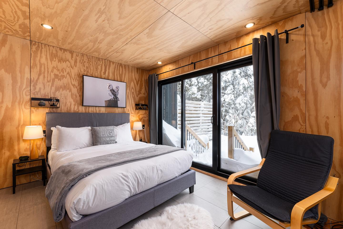 Chambre moderne en bois avec lit double, fauteuil et vue enneigée par les baies vitrées.