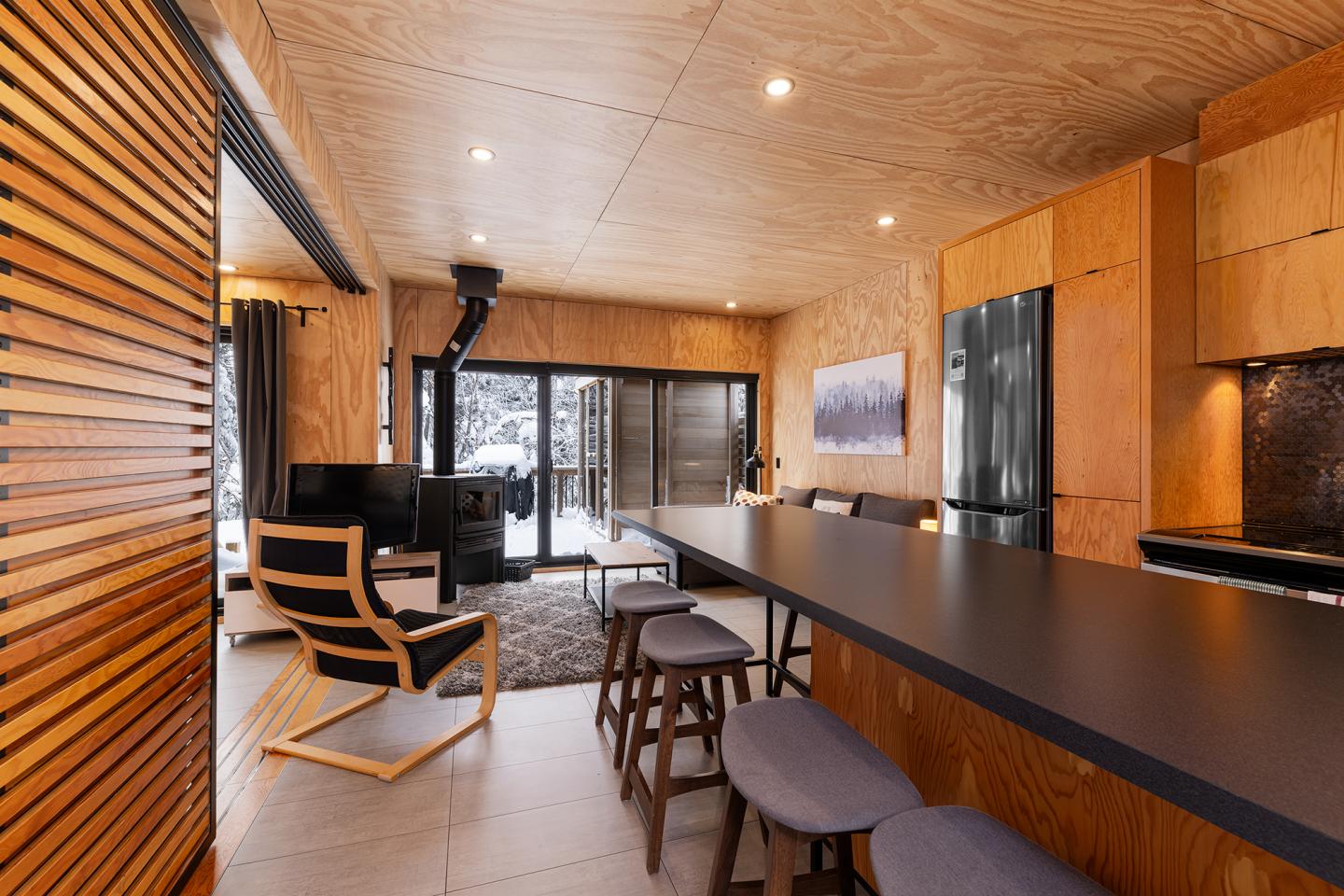 Salon moderne en bois avec cuisine ouverte, grande table et cheminée, vue sur la neige.