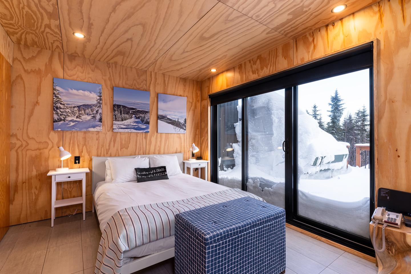 Chambre moderne en bois avec vue sur neige, lit blanc et lampes.