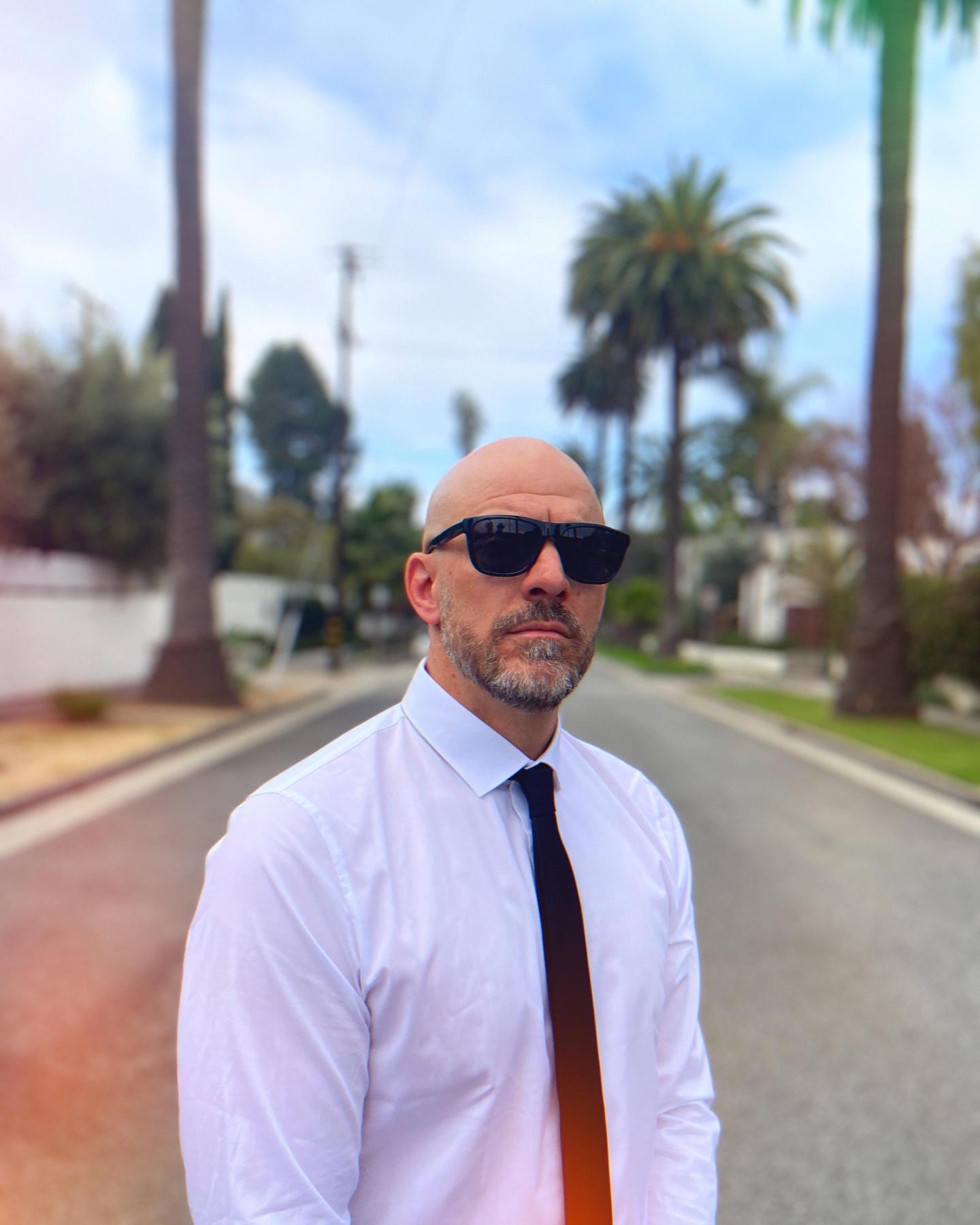 Homme en chemise blanche et cravate noire dans une rue bordée de palmiers.