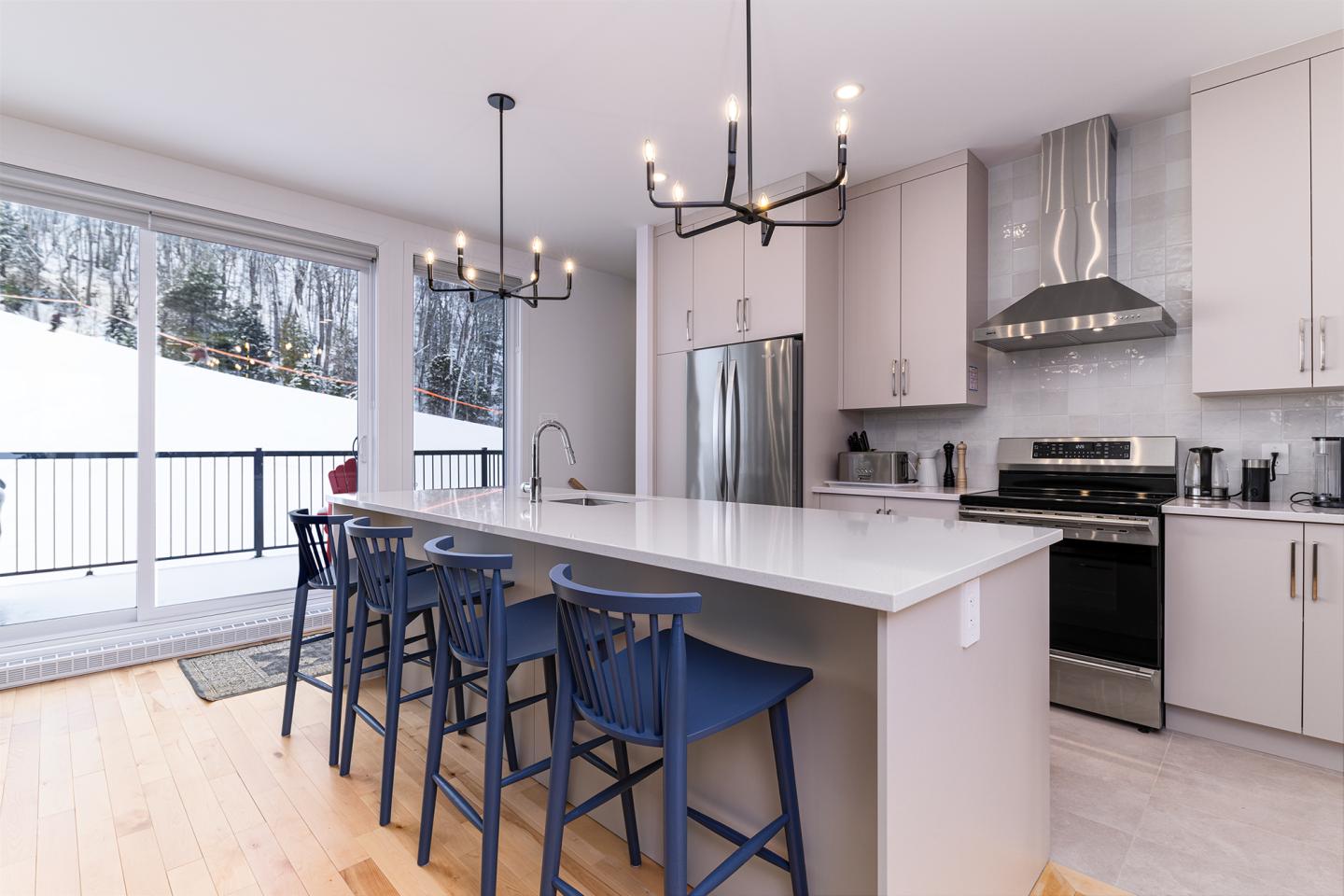 Cuisine moderne avec îlot, chaises bleues, et grand réfrigérateur. Fenêtre sur paysage enneigé.