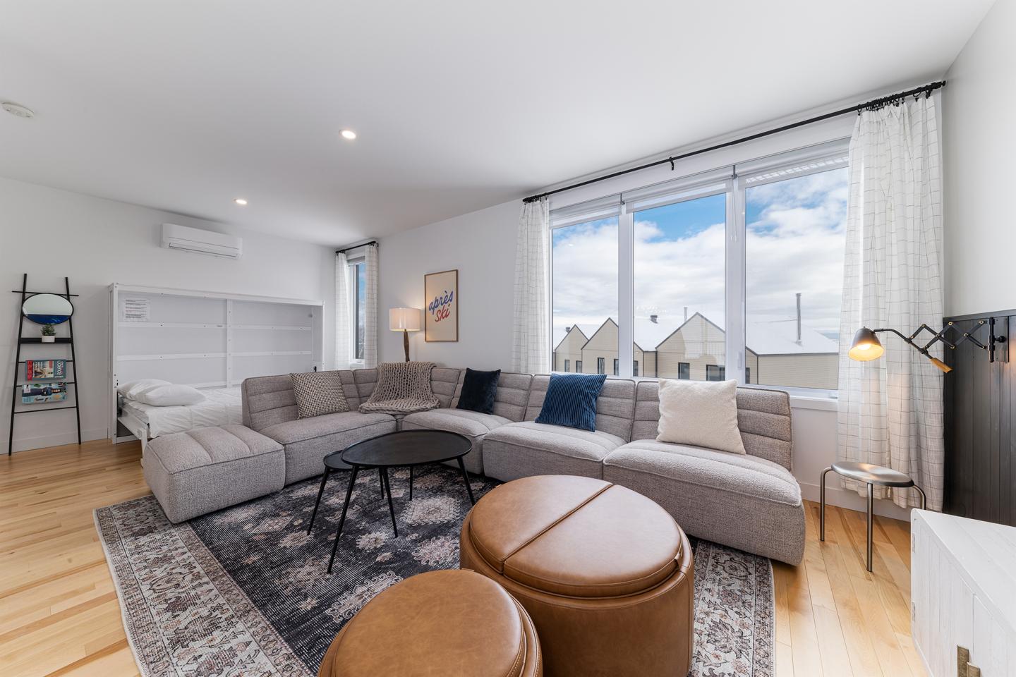 Salon moderne lumineux avec canapé gris et poufs marron, grandes fenêtres.