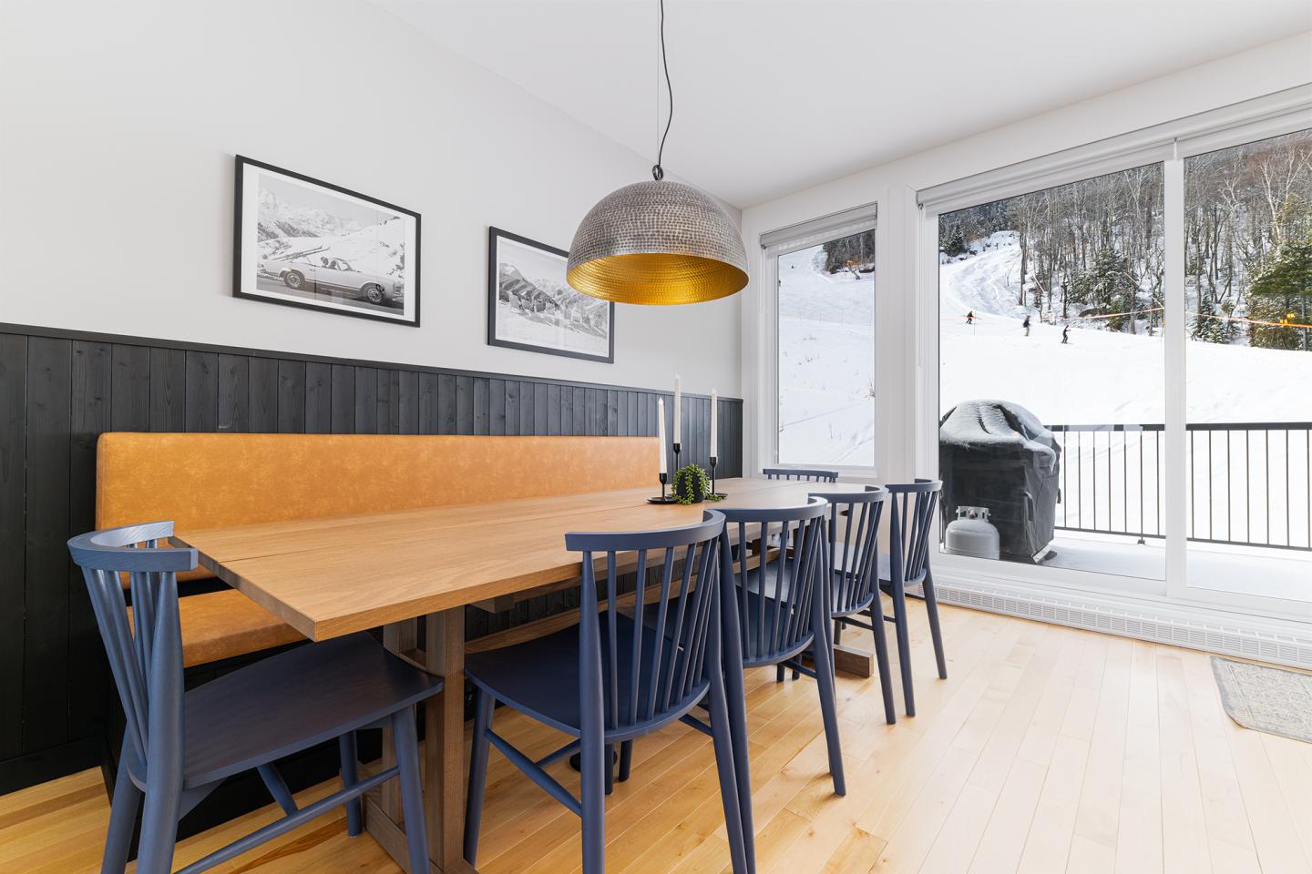 Salle à manger avec table en bois, chaises bleues, et grandes fenêtres donnant sur neige.