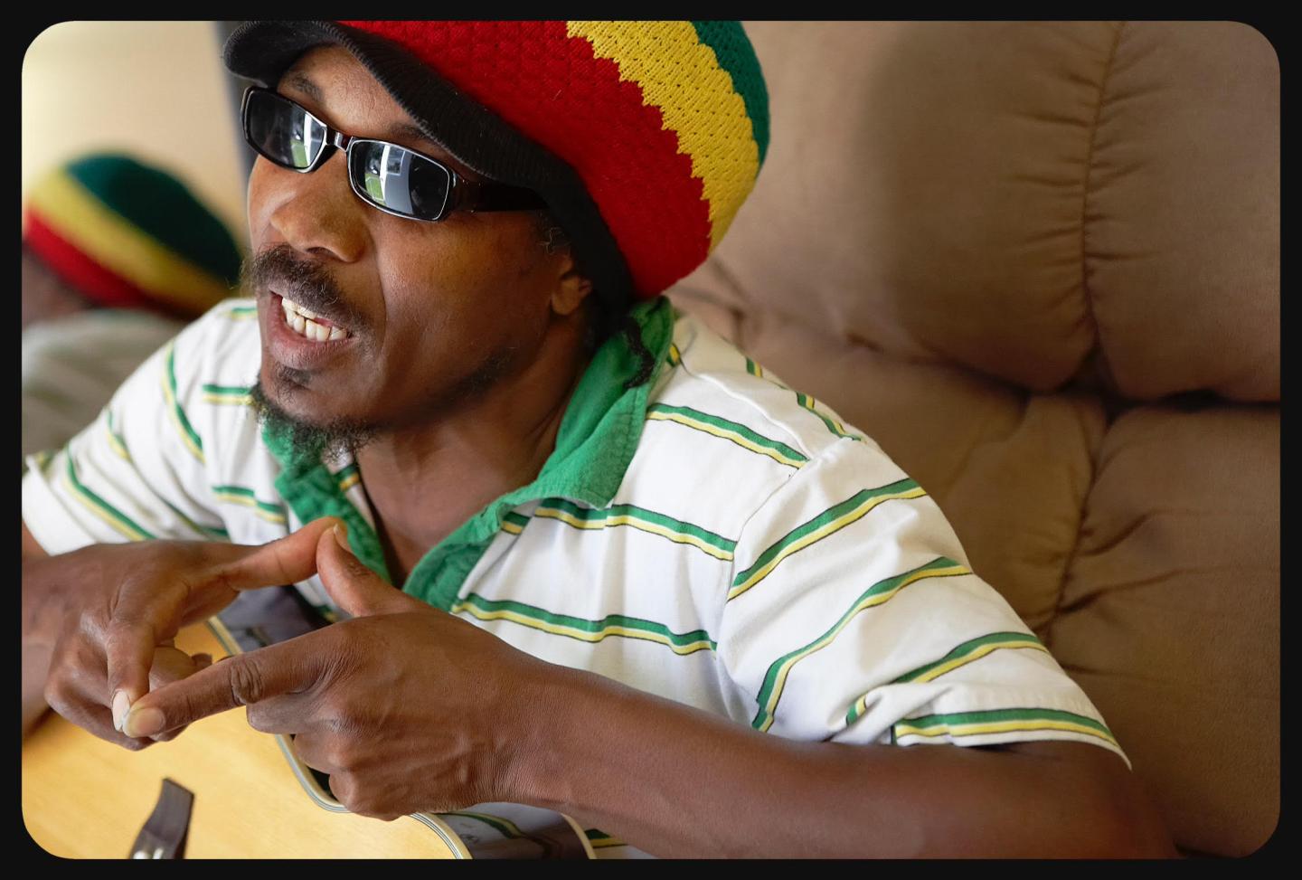 Homme avec lunettes, chapeau multicolore, joue de la guitare sur un canapé.