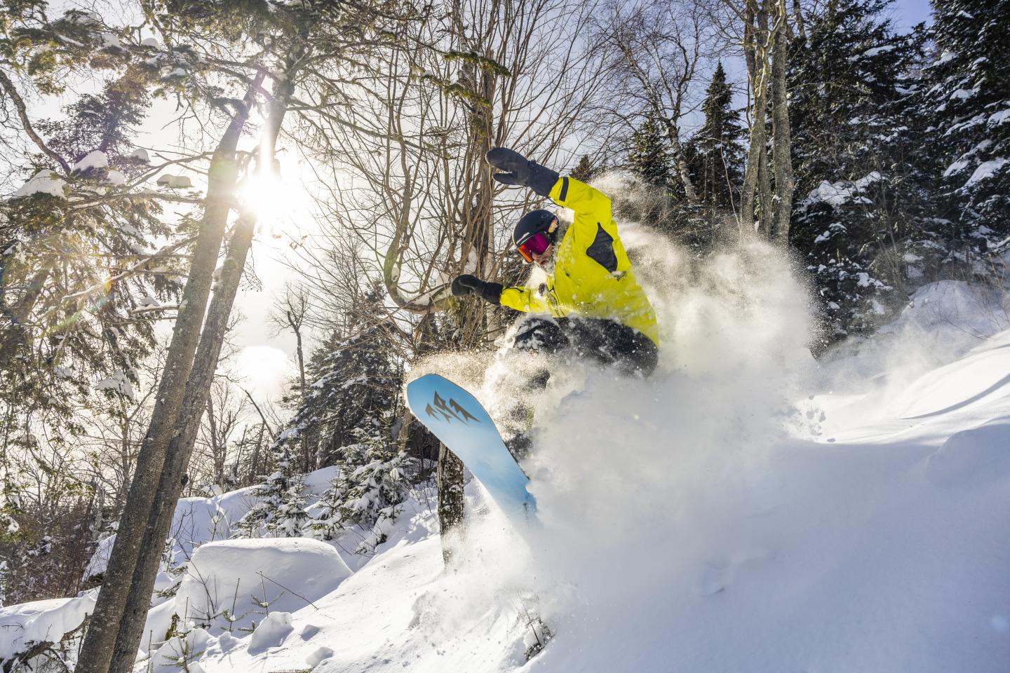 Snowboardeur en descente dans une forêt enneigée, soleil éclatant.