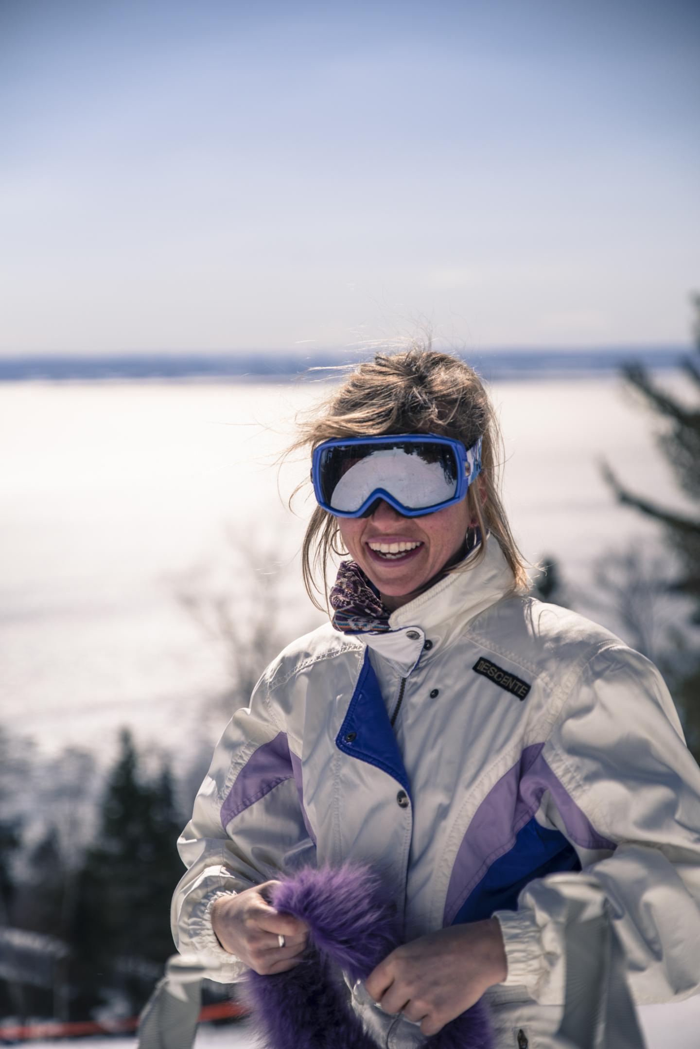 Femme souriante en tenue de ski, montagnes enneigées en arrière-plan.