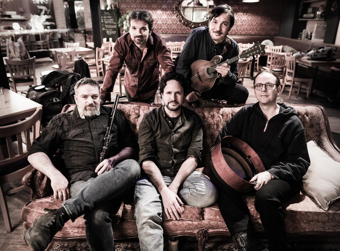Cinq musiciens assis dans un café chaleureux, souriant. Un joueur de mandoline debout derrière.