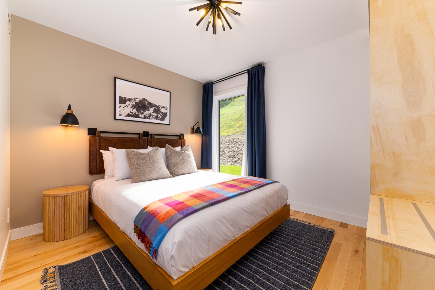 Chambre lumineuse avec lit double, plaid coloré, et grande fenêtre.