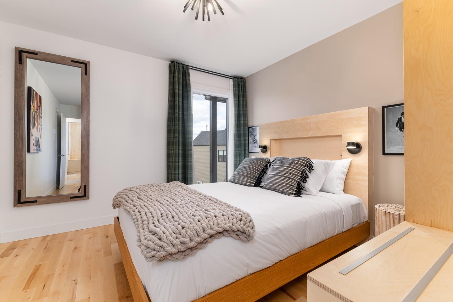 Chambre moderne avec lit double, couverture beige, miroir et grandes fenêtres.