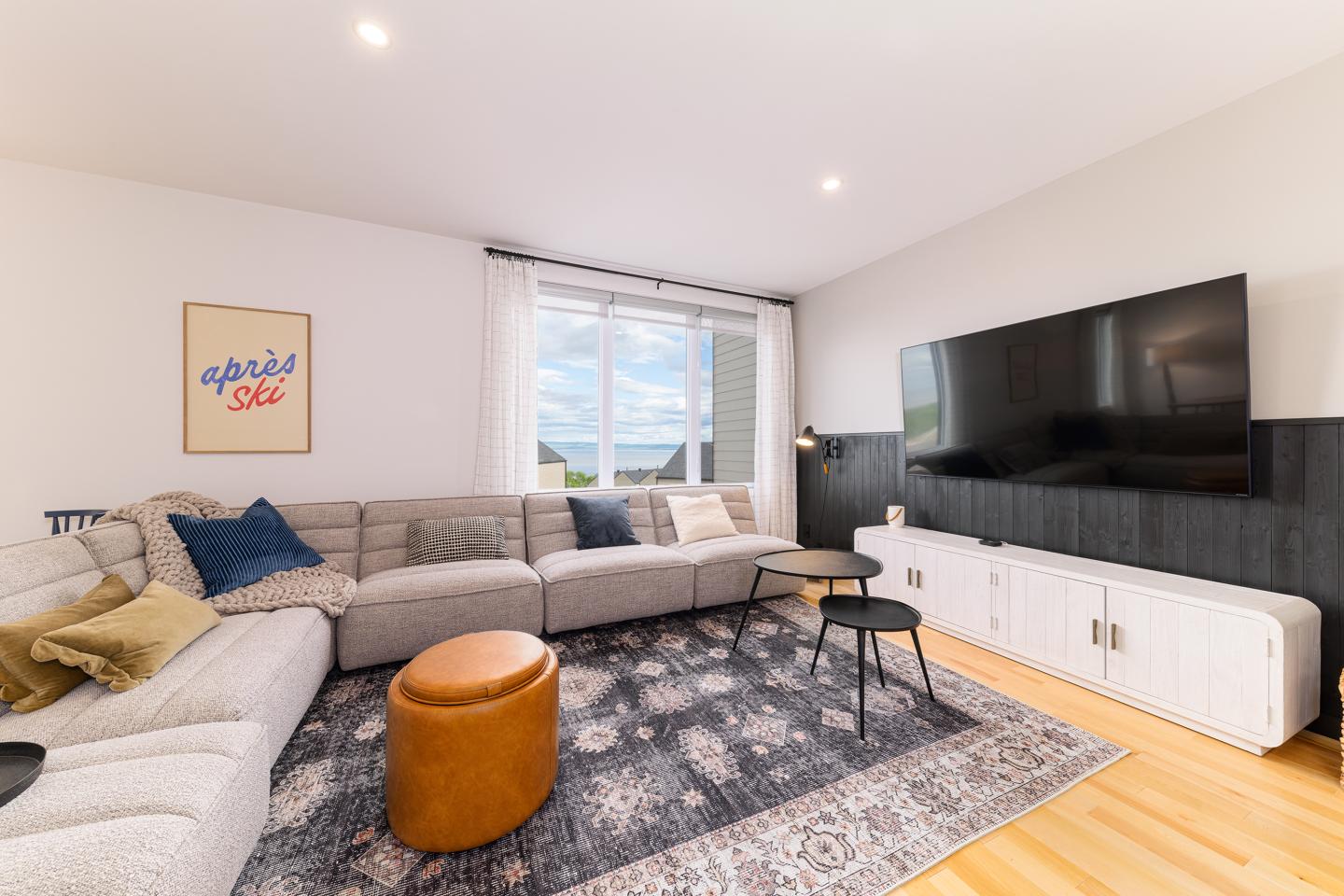 Salon moderne avec canapé gris, télé murale et table basse sur tapis.