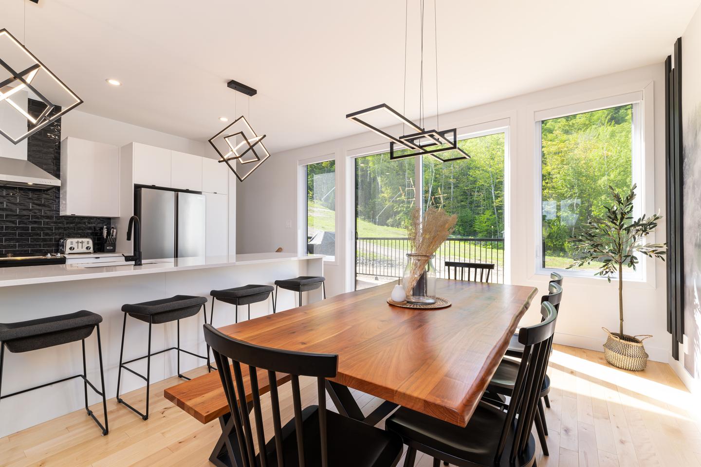 Salle à manger lumineuse avec table en bois, chaises noires et cuisine moderne à l'arrière.