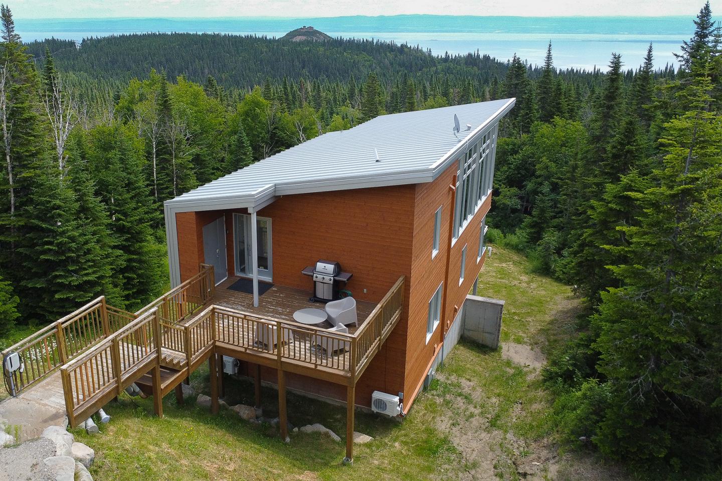 Maison en bois rouge avec terrasse, entourée de forêt, vue sur mer.
