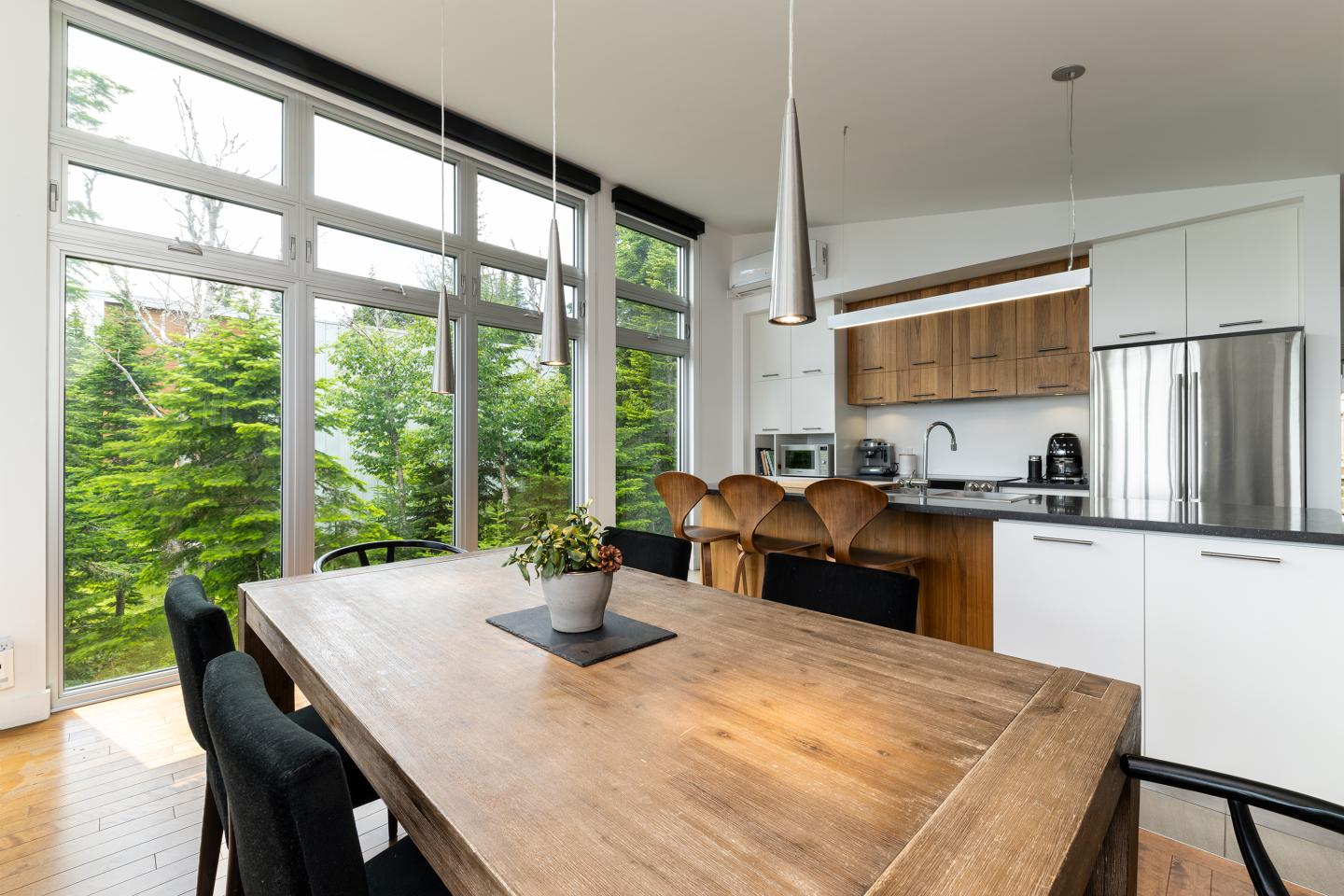 Cuisine moderne avec table en bois, grandes fenêtres, et plantes vertes à l'extérieur.