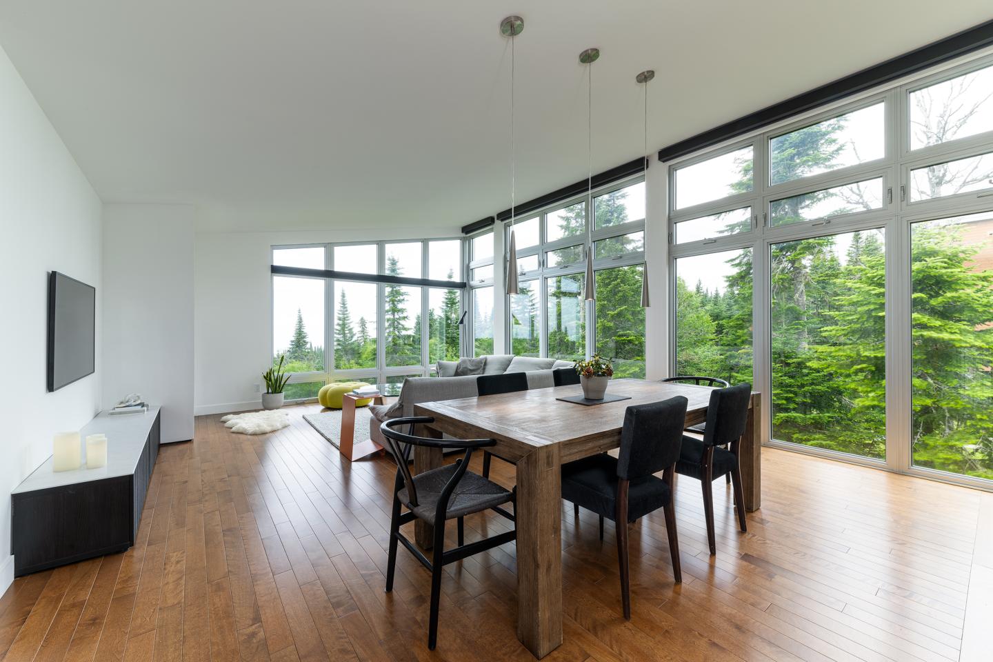 Salle à manger moderne avec de grandes fenêtres, table en bois, chaises noires, vue sur verdure.