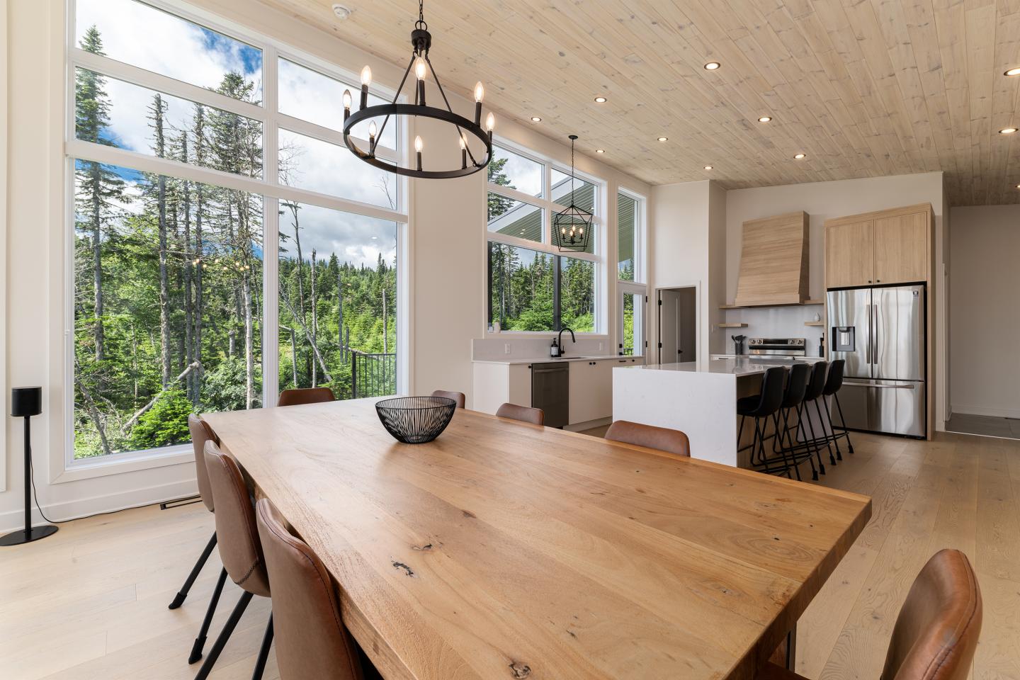 Salle à manger lumineuse avec grande table en bois et vue sur forêt par de larges fenêtres.
