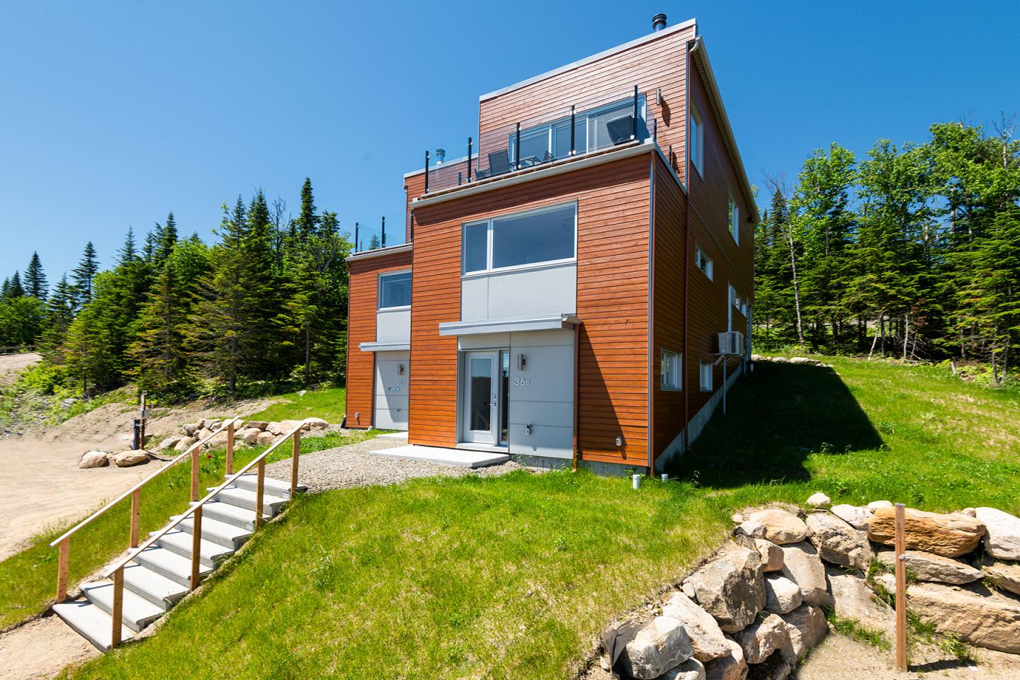 Maison moderne avec façade en bois, entourée de pelouse et de forêt sous un ciel bleu.