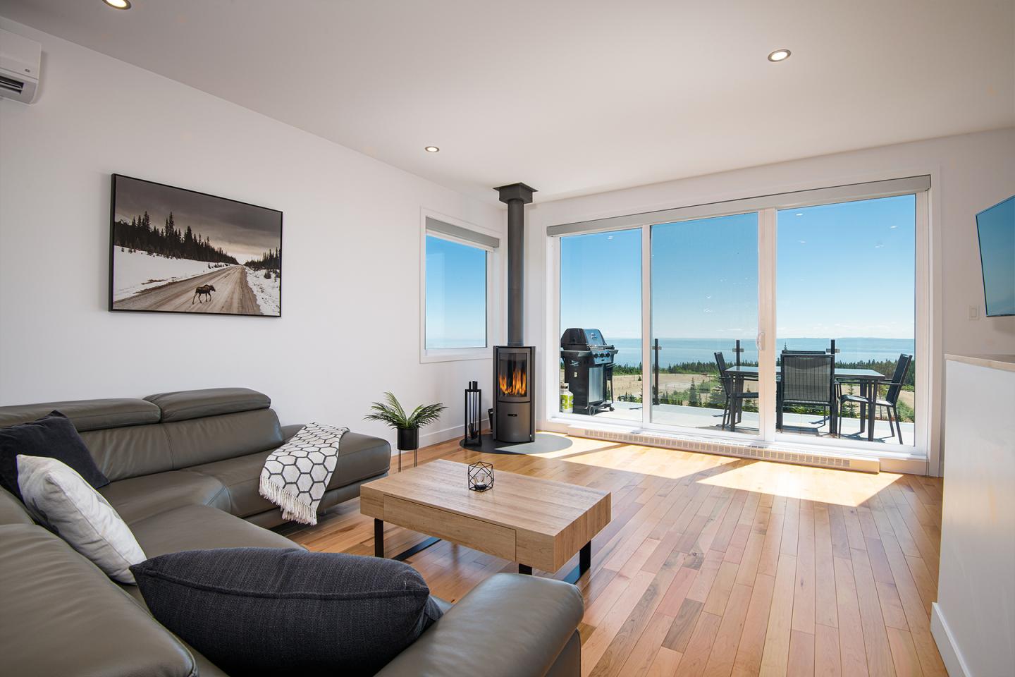 Salon moderne avec vue sur mer, canapé gris et poêle à bois.