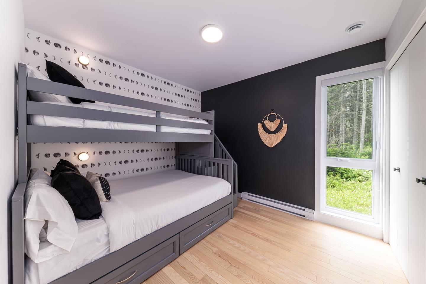 Chambre moderne avec lits superposés gris, coussins noirs, mur noir et vue sur jardin.