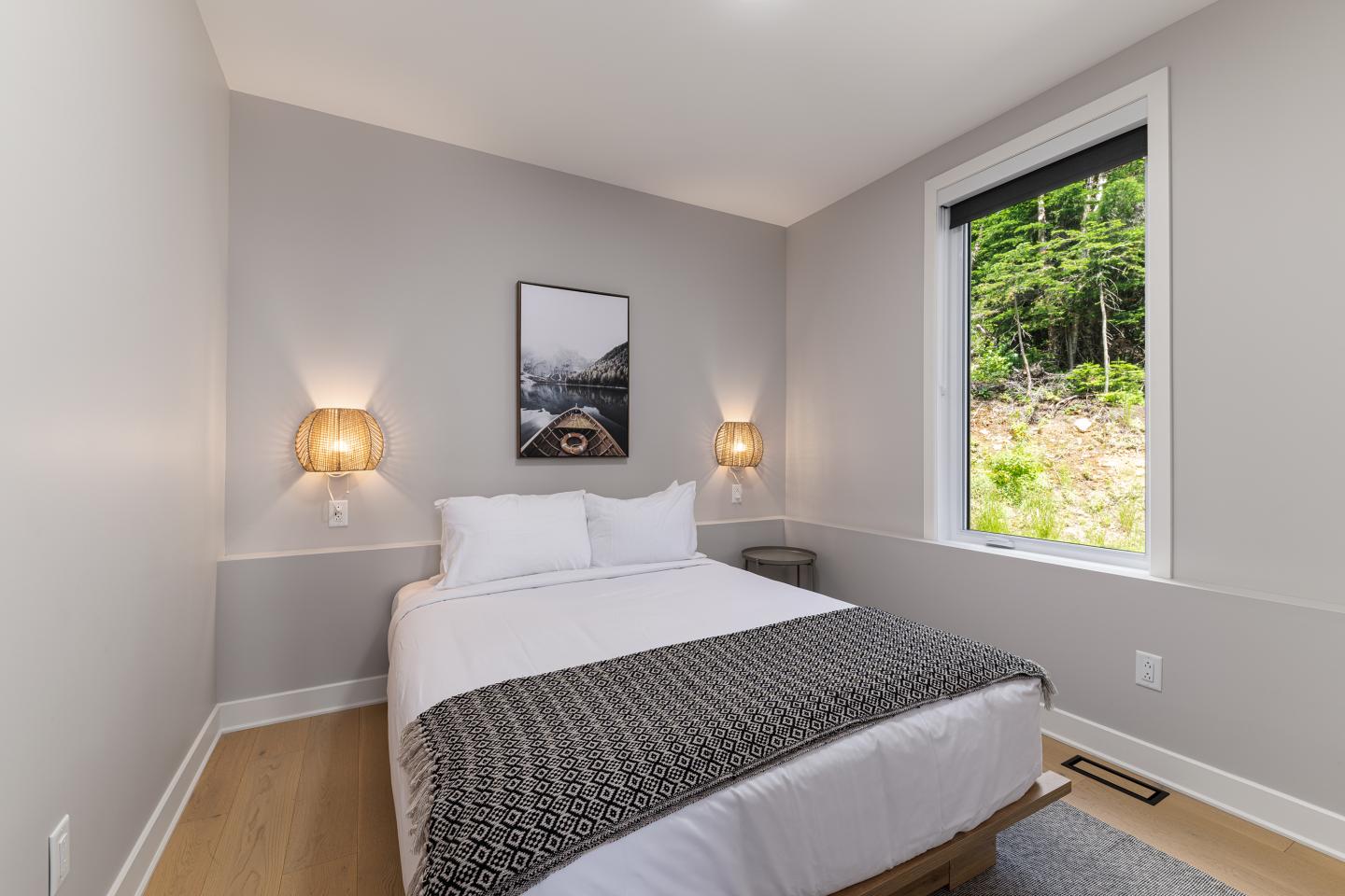 Petite chambre moderne avec lit double, deux lampes murales et fenêtre avec vue sur la forêt.