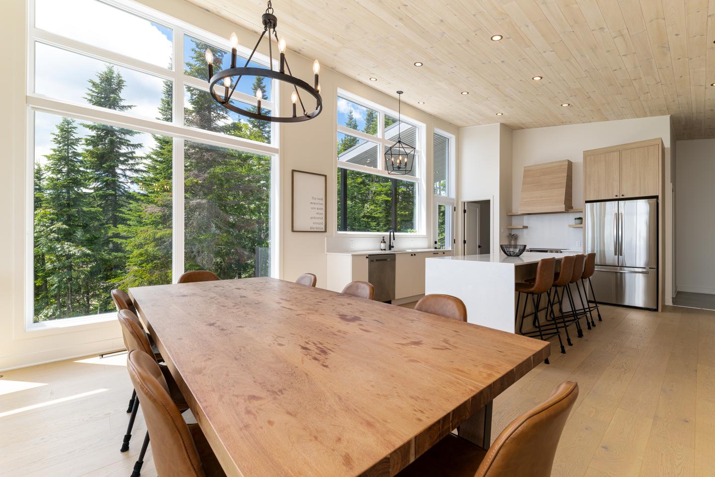 Salle à manger lumineuse avec grande table en bois, chaises marron, et cuisine moderne à l'arrière.