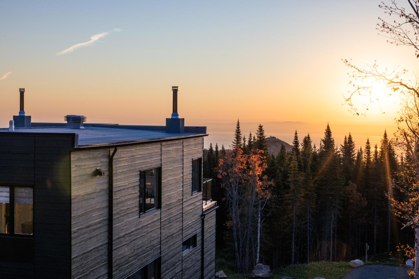 Maison moderne en bois au coucher du soleil, entourée d'arbres et vue sur l'horizon.