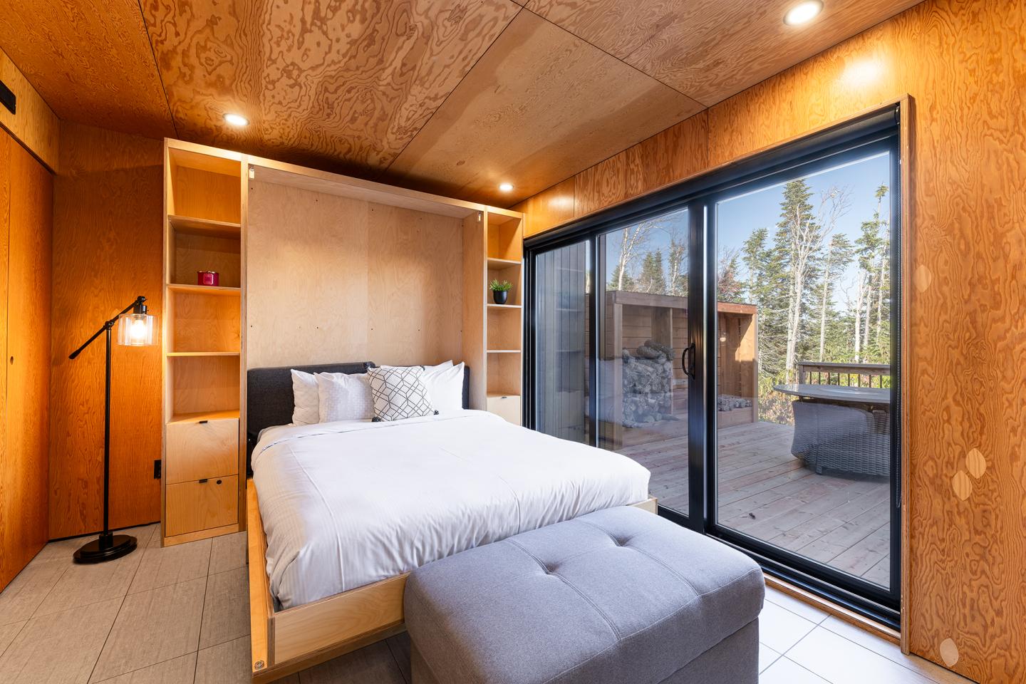 Chambre moderne en bois avec lit double, grande baie vitrée sur terrasse et forêt.