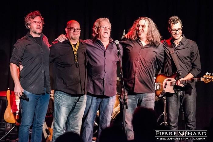 Cinq musiciens sur scène, souriants, bras autour des épaules, guitares à gauche et droite.