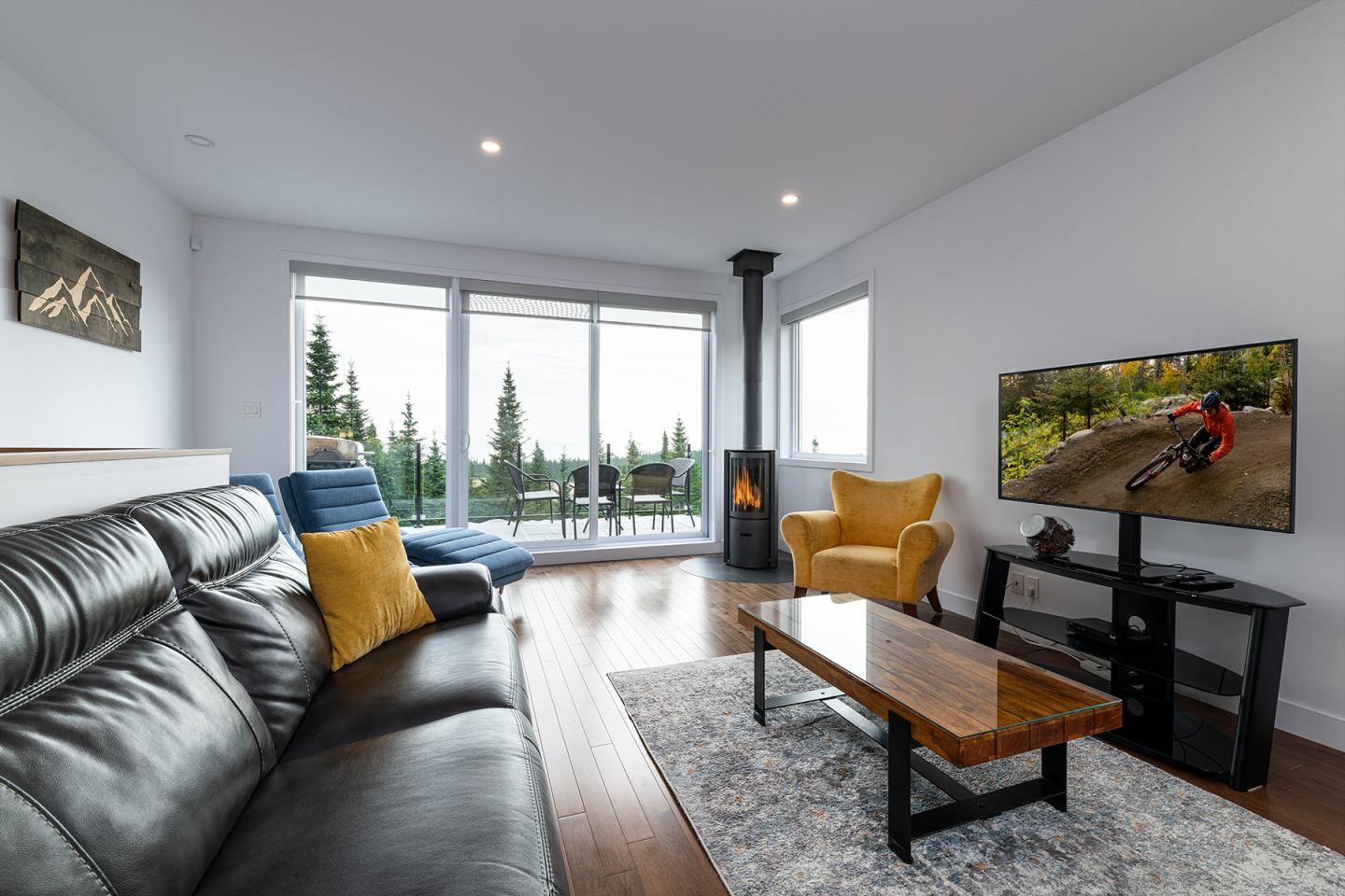 Salon moderne avec canapé en cuir, télé allumée et vue sur des arbres par la baie vitrée.