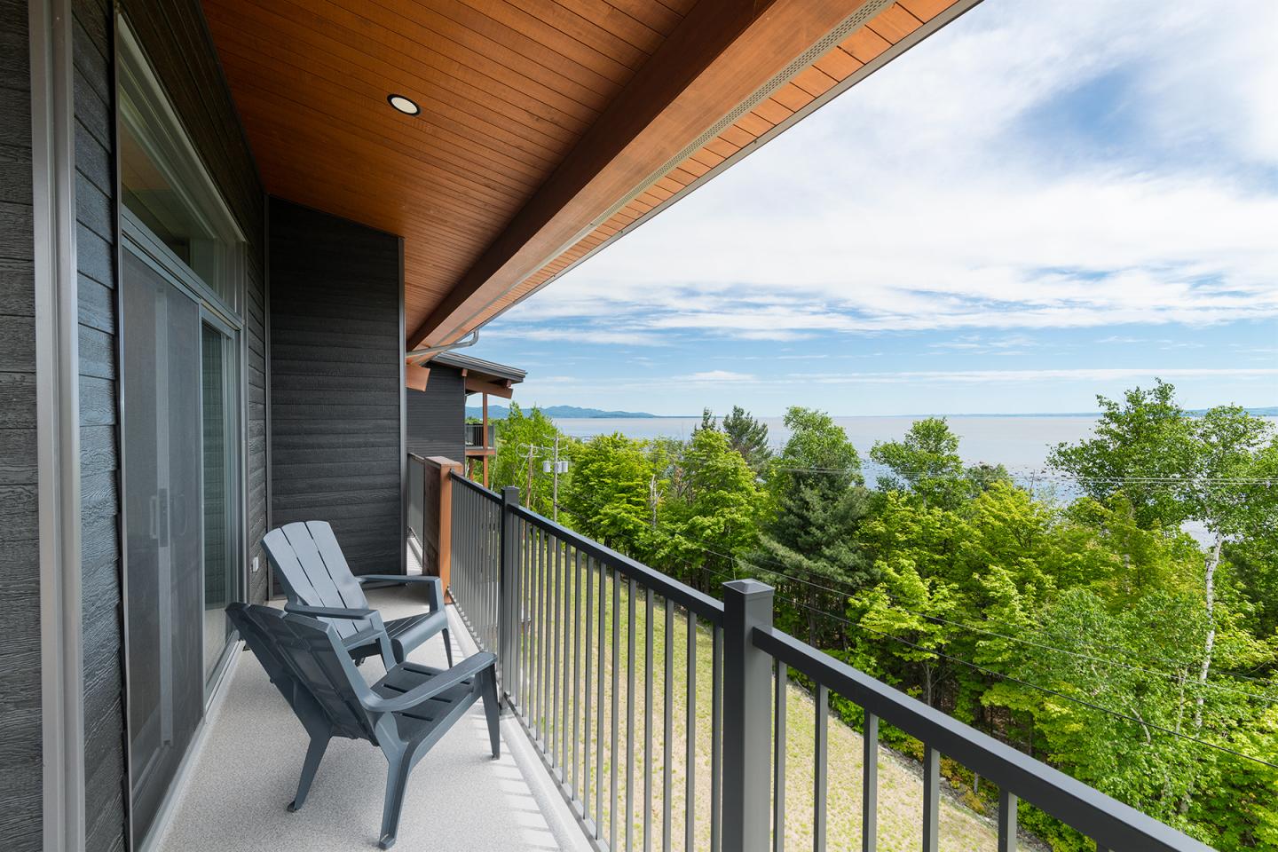 Balcon avec chaises en bois, vue sur arbres et mer au loin.