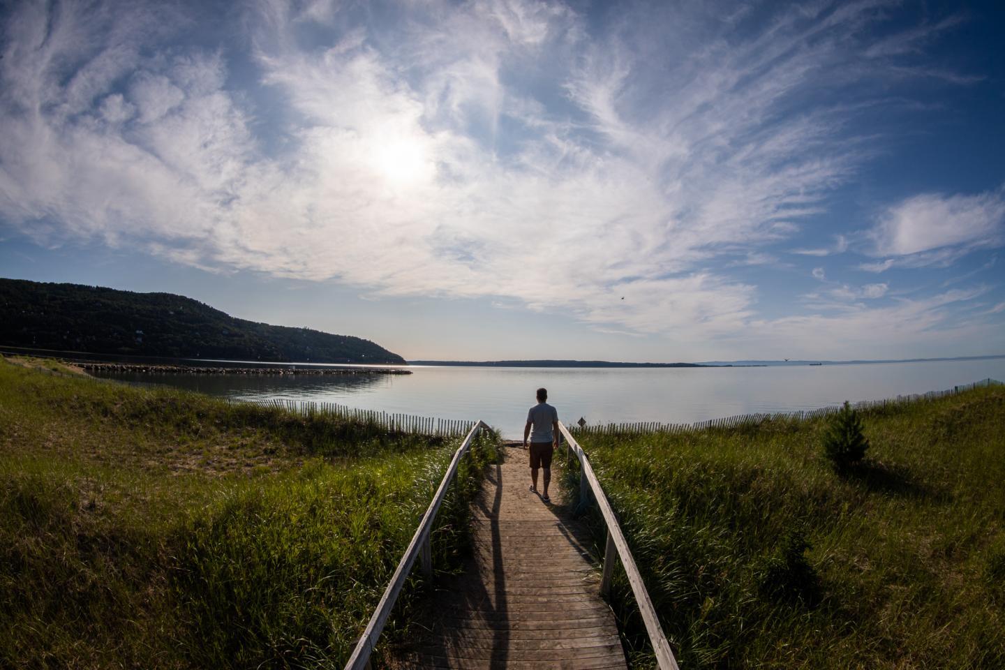 Personne marchant sur une passerelle vers un lac, ciel nuageux et montagnes en arrière-plan.