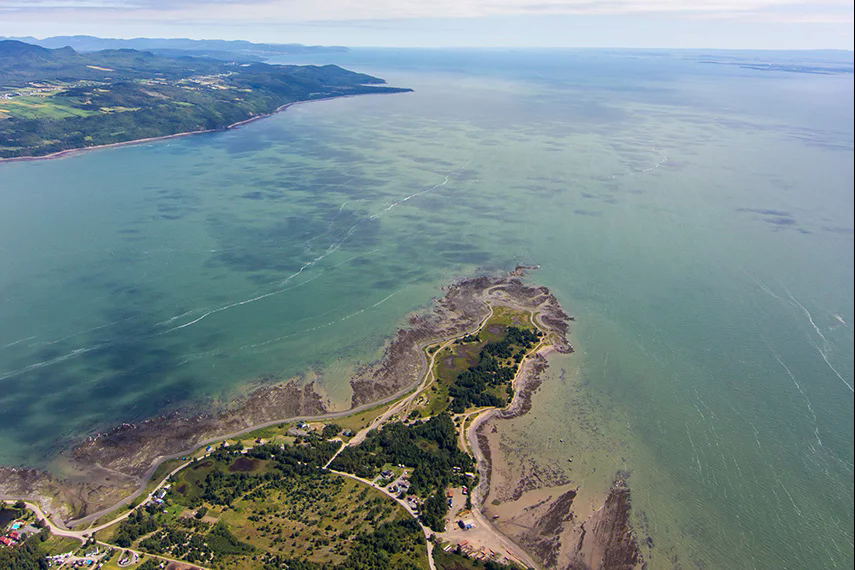 Vue aérienne d'un littoral avec mer turquoise, végétation verte et ciel dégagé.