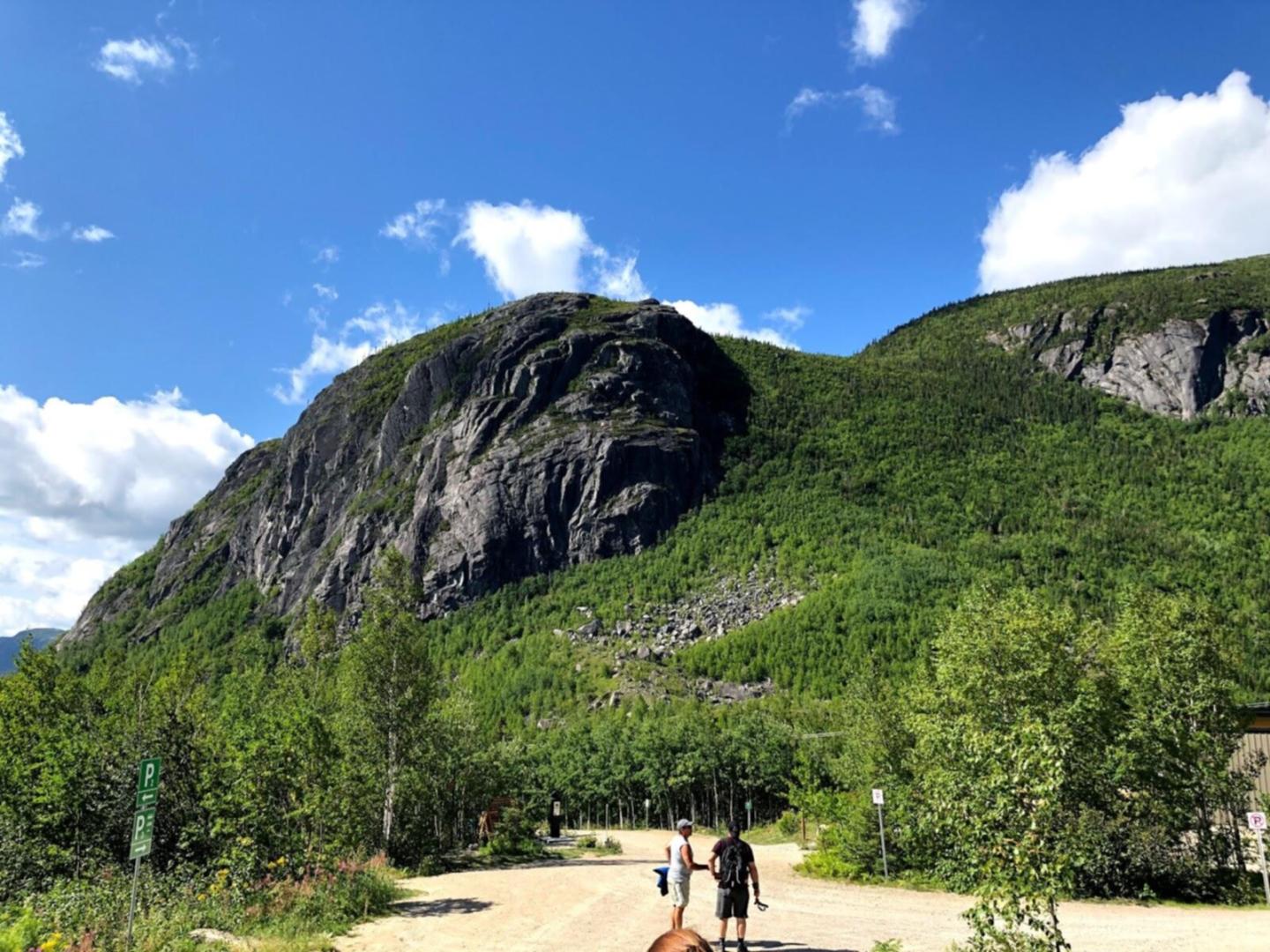 Montagne verdoyante sous un ciel bleu avec quelques nuages, personnes marchant sur un sentier.