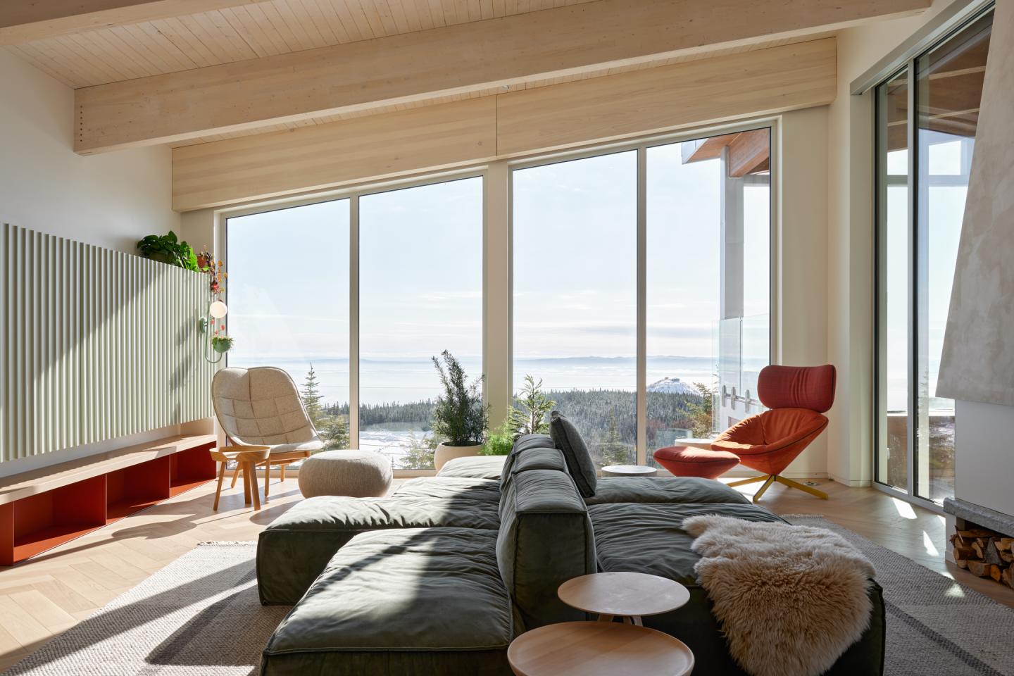 Salon lumineux avec grandes fenêtres, canapé vert, fauteuil rouge, vue sur la mer.