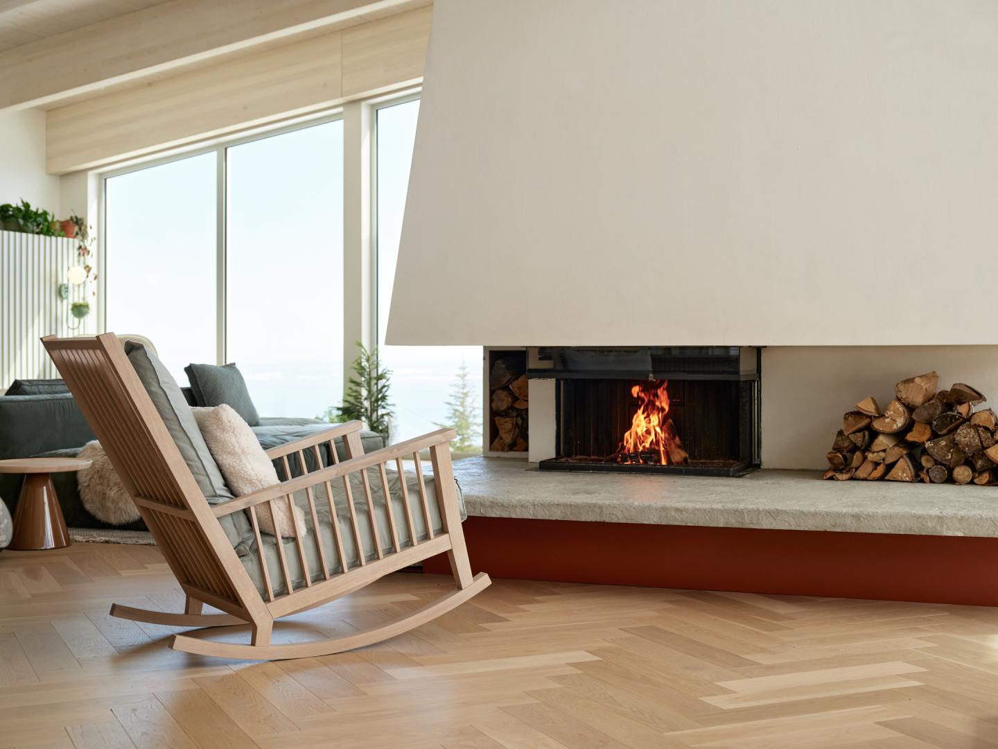Chaise berçante en bois près d'une cheminée allumée dans un salon lumineux.