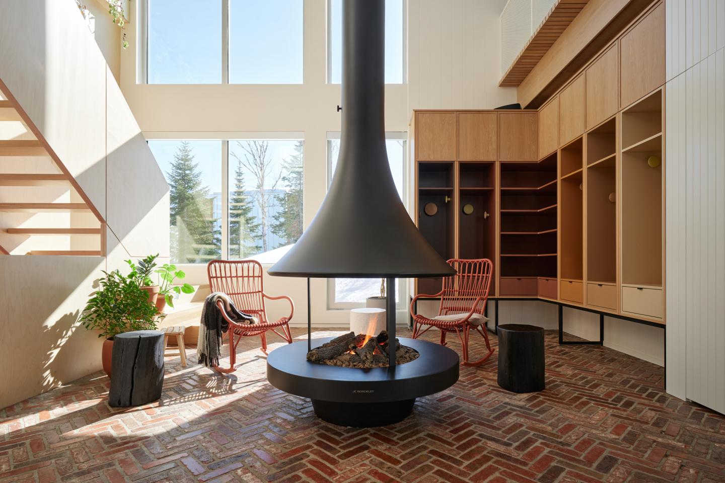 Salon moderne avec cheminée centrale, sol en briques, grandes fenêtres et fauteuils rouges.