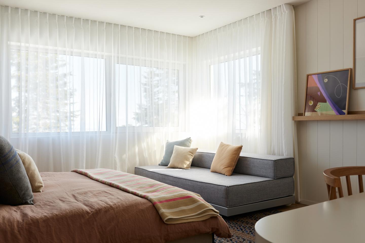 Chambre lumineuse avec lit et canapé gris, rideaux blancs et tableau coloré.