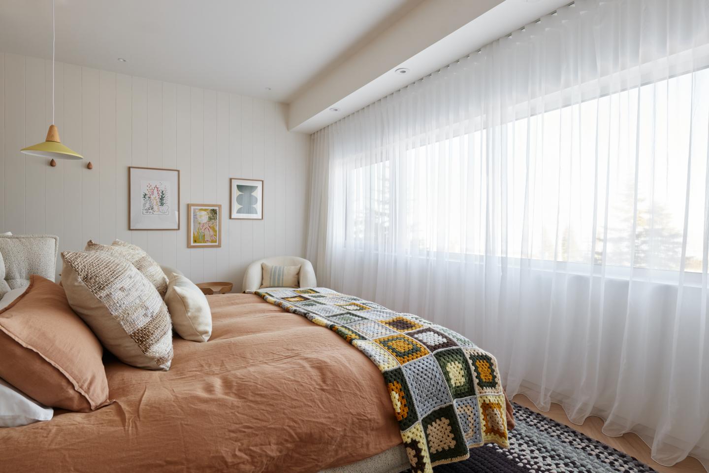 Chambre lumineuse avec lit beige, couverture colorée et grandes fenêtres voilées blanches.