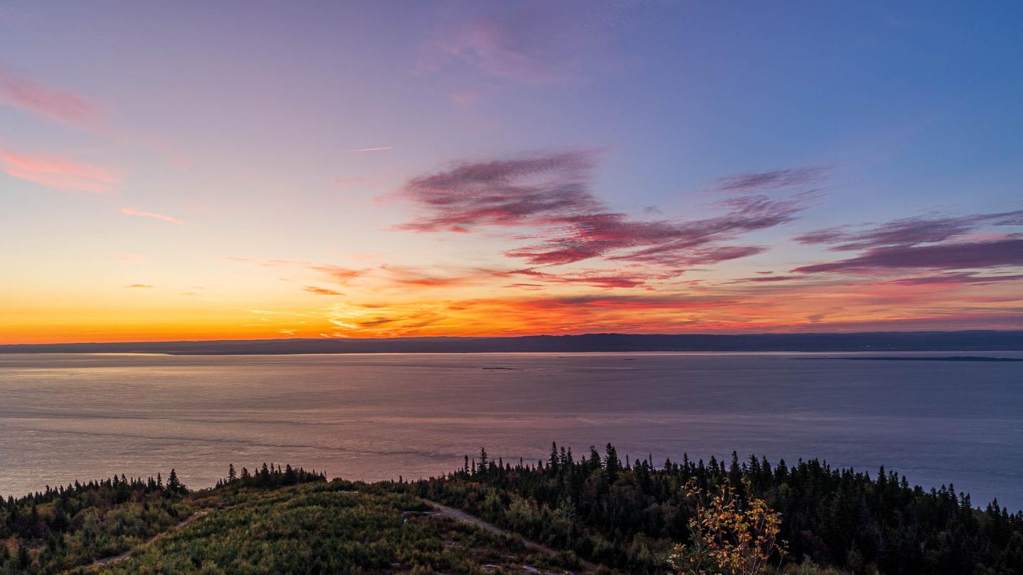 Coucher de soleil sur un lac avec ciel coloré et forêt au premier plan.